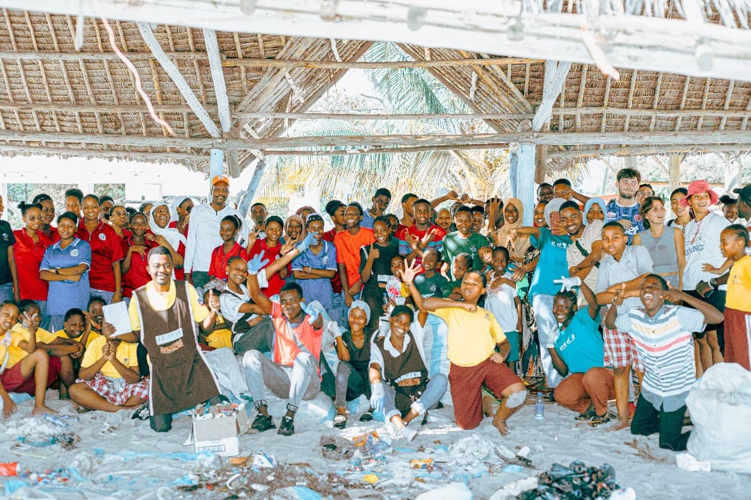 A large group of volunteers under a thatched shelter with sorted waste in front of them